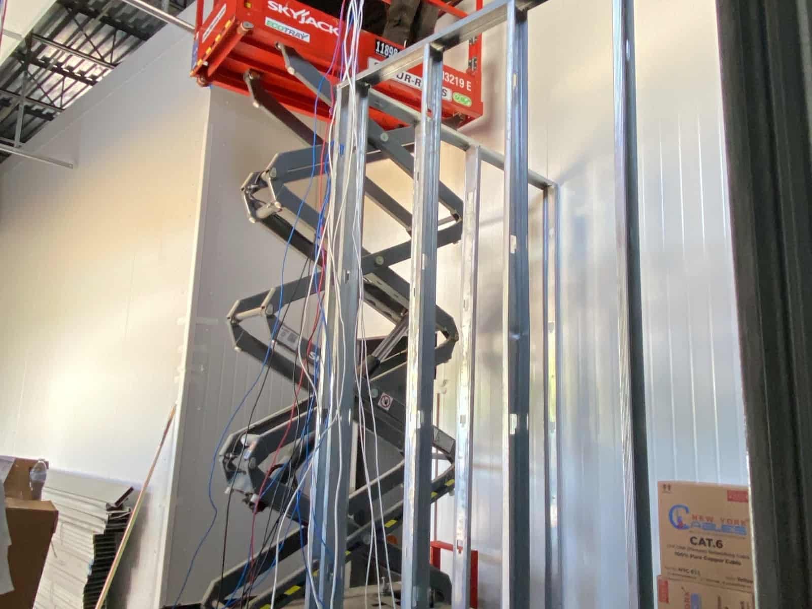 Technician on a scissor lift pulling Cat6 cable through steel stud framing, with boxes of Cat6 stacked below