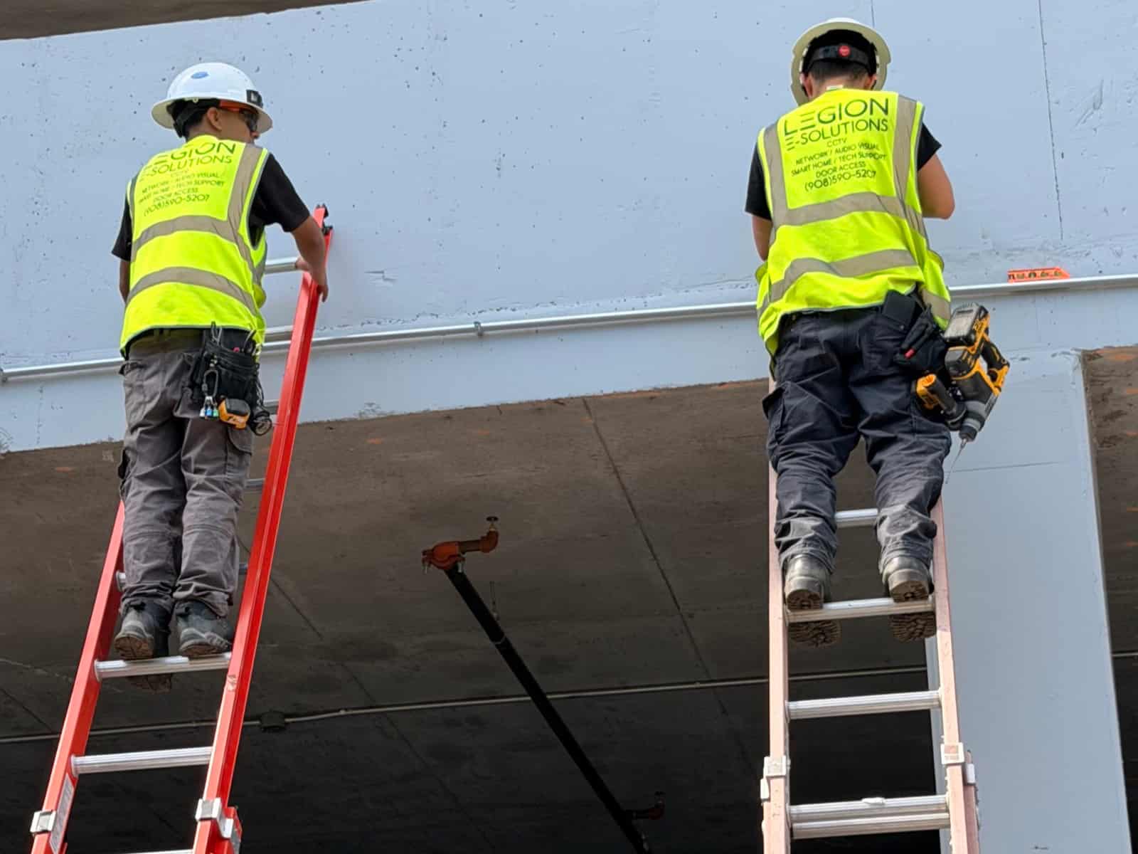 Two Legion E-Solutions technicians on ladders running conduit along a concrete parking deck ceiling