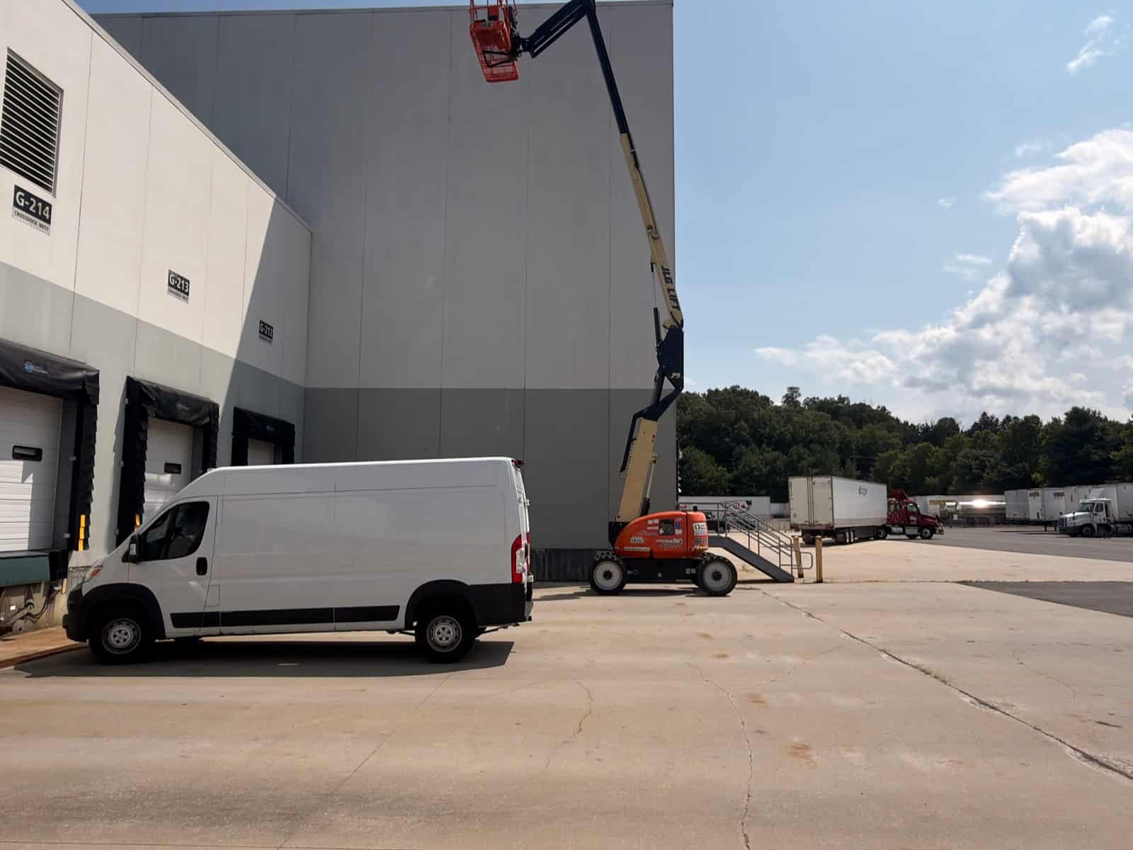 Boom lift raised against a distribution warehouse wall beside loading docks, with a work van parked below